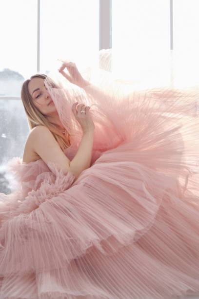 a girl in a beautiful lush long dress of dusty rose color stands near an open window. vintage bridal glamor. fashion shooting in a wedding or evening dress - clothing fashion model old fashioned women imagens e fotografias de stock