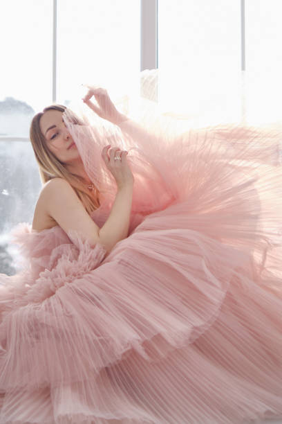 девушка в красивом пышном длинном платье пыльного розового цвета стоит возле открытого окна. винтажный свадебный гламур. модная съемка в с� - floral модель стоковые фото и изображения