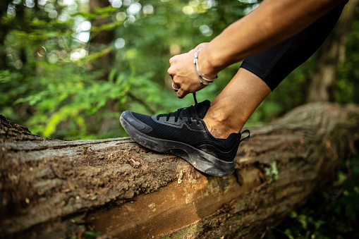 Unrecognizable female tying black running shoes, while foot is on a log in forest
