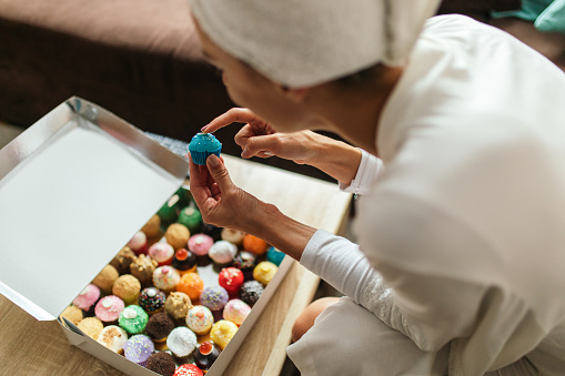 Young woman is very happy when opening package box with lot of multicolored cakes purchased online that she just received. She is wearing bathrobe and her hair is wrapped in towel while sitting in her living room. Online shopping and subscription services concept