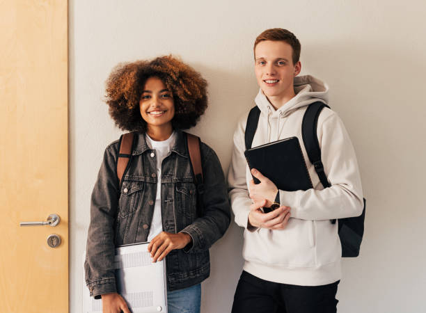 zwei lächelnde klassenkameraden, die zusammen an der wand stehen und in die kamera schauen - highschool student stock-fotos und bilder