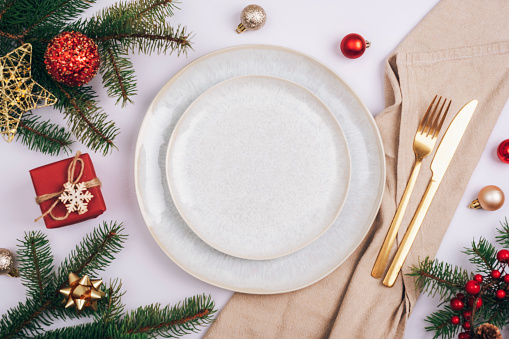 Stylish table setting, fir branches, stars and balls with white plate, cutlery and beige napkin on white table. Christmas concept. Top view, flat lay.