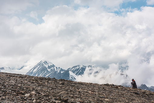 Hiker and high snowy mountains in dense low clouds. Minimal scenery with man on stone hill against beautiful mountain peaks in thick clouds. Simple minimalism with man and majestic mountains nature.