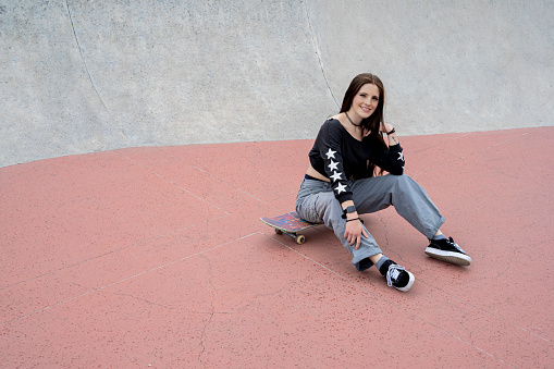 Young latina millennial girl looking at the camera sitting on a skateboard in a skatepark in the urban environment of the city of bogotá. Lifestyle and skate tricks.
