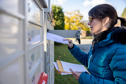 A woman is collecting post at home in autumn, Quebec, Canada. She is smiling and unlocking the mailbox. She is pickup-up her mail and looking at the letters she received.