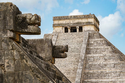View of El Castillo (Temple of Kukulkan) behind carved jaguar heads. Blue sky and clouds in the far background.