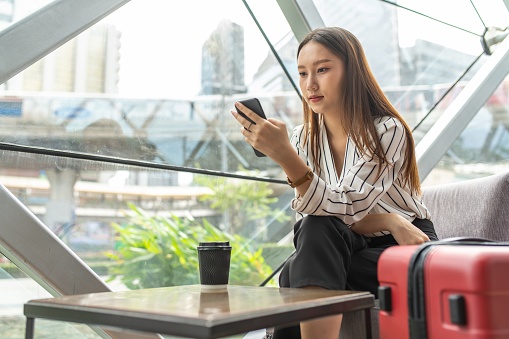 Portrait photo of young asian female solo lady traveller watching content on her smartphone while waiting for departure in a cafe in airport terminal