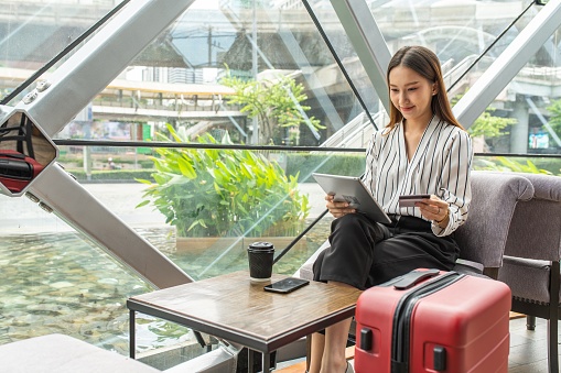 Portrait photo of young asian female solo lady traveller doing online shopping and payment on her digital tablet while waiting for departure in a cafe in airport terminal