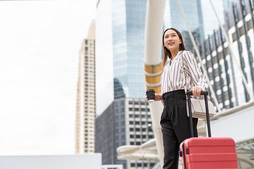 Portrait photo of a young asian smart casual business lady holding a cup of coffee with her red luggage on the way to a business trip from her office