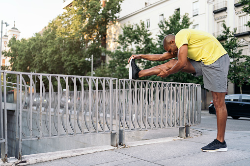 African American man stretching his right leg over railing in the city,