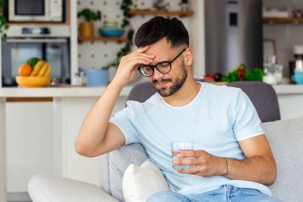 young man suffering from strong headache or migraine sitting with glass of water on the sofa, millennial guy feeling intoxication and pain touching aching head, morning after hangover concept - intoxication imagens e fotografias de stock