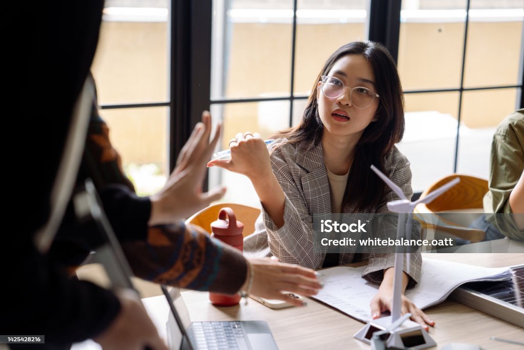 Young business team developing renewable energy project. Diverse team of engineers sitting at office table while talking and creating renewable energy project, using blueprints, solar panel and windmill models. Manufacturing Stock Photo