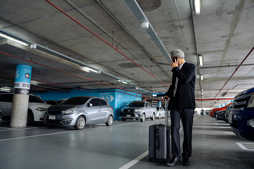 Asian senior businessman talking on the phone in airport parking lot with happy face while talking on the phone he looked at his wristwatch so that he can get on the plane in time to travel to talk business abroad.