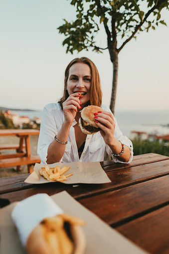 Happy woman sitting outdoors and eating burger and fries on a sunny summer day during a seaside vacation