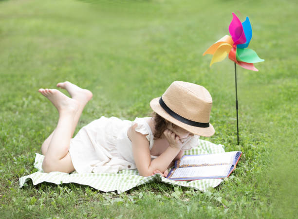 Cute little girl in a hat lies on a blanket and reads a book at a picnic in nature stock photo