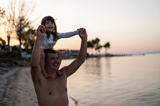 A man and his daughter are walking on the beach, and the girl is sitting on her father's shoulders.