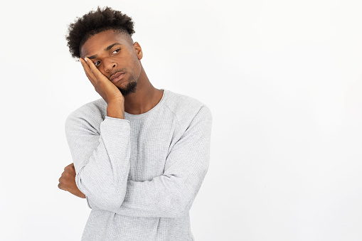 Portrait of bored African American man looking away. Young bearded guy wearing white sweater standing leaning on hand against white background. Boredom and depression concept