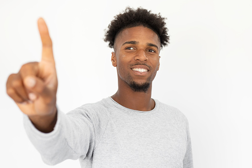 Portrait of happy African American man touching invisible wall. Young bearded guy wearing white sweater pointing at camera and smiling against white background. Interactive technology concept