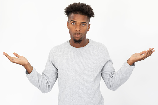 Portrait of ignorant African American man shrugging shoulders. Young bearded guy wearing white sweater looking at camera in confusion against white background. Ignorance and uncertainly concept