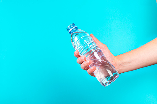 Front view of a young man hand holding a bluish translucent PET  water bottle against a blue background. The hand is at the upper right corner leaving a useful copy space at the left side of the image.