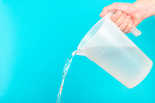 Front view of a young man hand holding a polypropylene white and opaque white pitcher and pouring water against a solid blue background