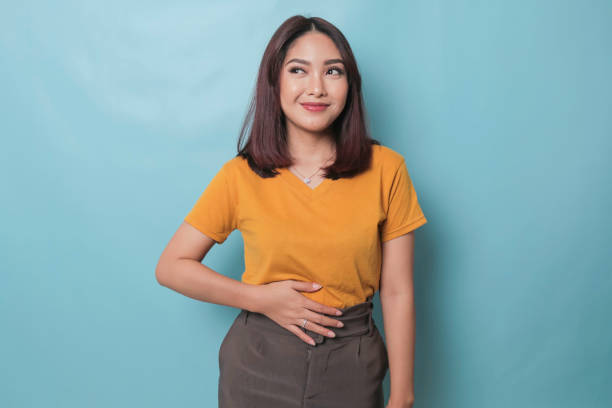 complacida y alegre mujer asiática mantiene la mano en el vientre se siente llena después de la deliciosa cena vestida casualmente de pie pensativo sobre fondo azul. - abdomen fotografías e imágenes de stock