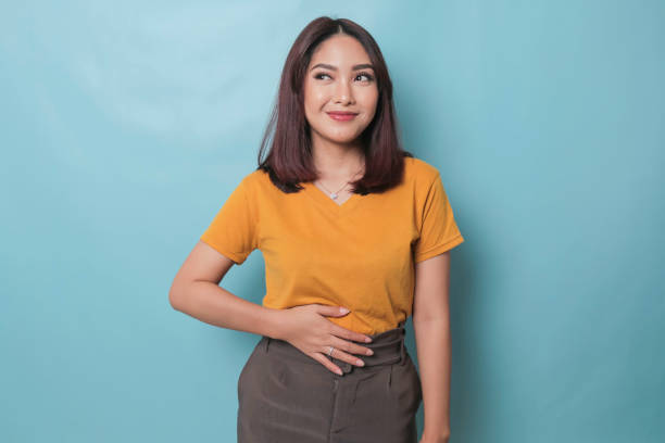 pleased cheerful asian woman keeps hand on belly feels full after delicious dinner dressed casually stands thoughtful against blue background. - buik stockfoto's en -beelden