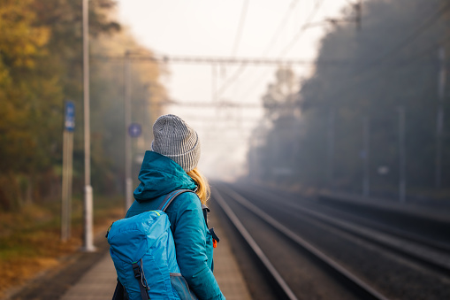 Female tourist with backpack waiting for train on empty railroad station alone. Woman travel at fall season