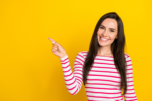 Photo portrait of stunning young woman toothy smile point offer empty space wear stylish striped outfit isolated on yellow color background.