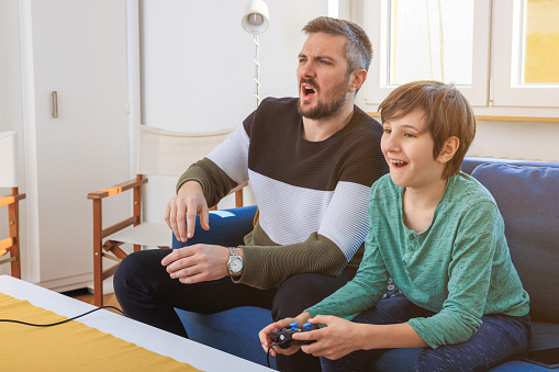 Happy boy playing video game with controller in hands, his father sitting next to him on the sofa and making a face
