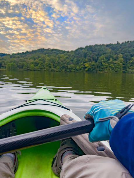 A personal perspective paddling Salt Fork Lake (State Park) in Ohio.   The unrecognizable person is holding the paddle and is wearing gloves, water shoes, and a blue sun shirt.  There are rolling hills in the distance. The season is summer.  The scene is early morning.