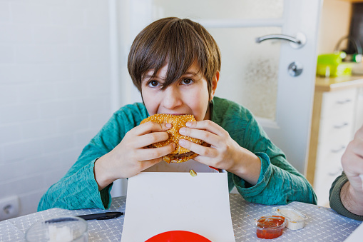 Boy sitting at the table at home and taking a bite of burger, holding it in hands, take out food delivery