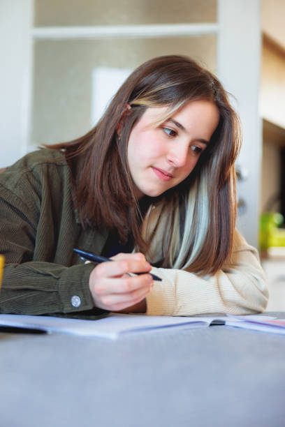 Teenage girl writing in her notebook at the table at home Teenage girl sitting at the table at home and doing school tasks in her notebook 2590 stock pictures, royalty-free photos & images