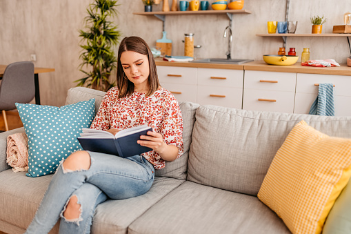 Beautiful young woman sitting on the sofa in the living room and reading a book.