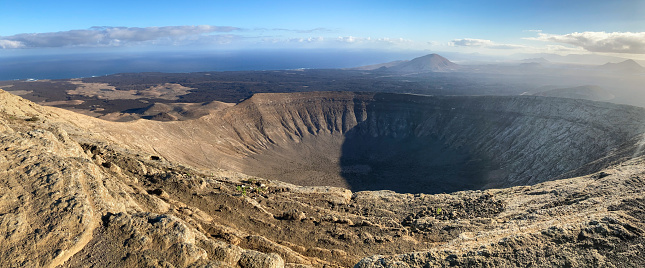 Panoramic image of the crater Caldera Blanca on early morning shot from the highest point Pico de la Caldera Blanca. In the background the volcano Montaña de Tenésara and the Atlantic Ocean. Lanzarote island, Spain.