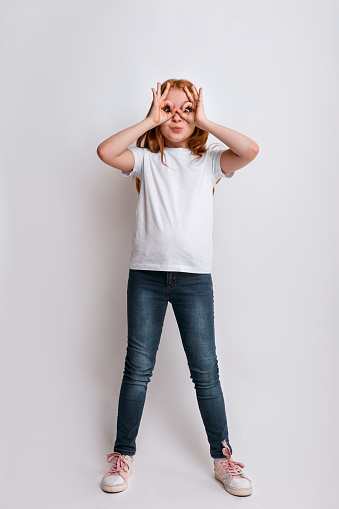 cheerful smiling redhead girl holding her hands near her eyes in the form of a mask wearing in white t-shirt on white background