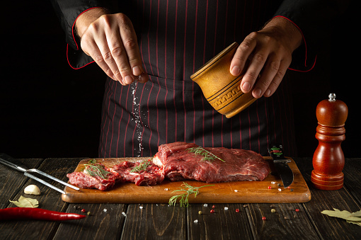 The chef salts raw fresh beef before roasting or grilling. Working environment in the kitchen of a restaurant or hotel.