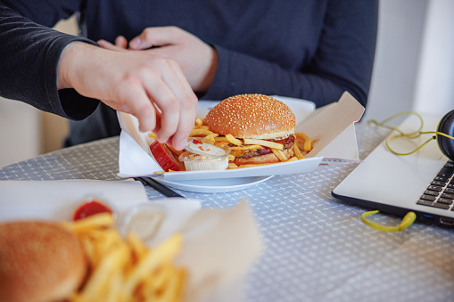 Eating burger and fries with dipping sauce at the table at home, taking a break from studies