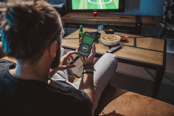 Watching soccer game at home Young man sitting on sofa at home and watching soccer game. He is using smart phone for sports betting . online soccer free betting stock pictures, royalty-free photos & images