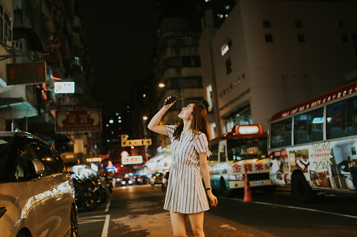 Young Asian woman taking photo of the view on the busy road with mini-busses and cars on the side, with a background of neon lights and local shops in the city. A happy Asian girl looking at the view and smiling while holding her camera and pointing it at the view upwards, in the background of Hong Kong at night. A portrait of a happy female traveler relaxing and discovering the streets of a buzzing city in the evening.