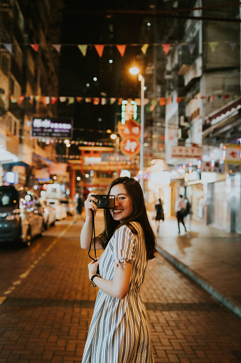 Young Asian woman holding her camera close to her face and looking backwards at the main camera on a street with neon lights and old buildings, in the city of Hong Kong. An Asian girl tourist using her camera to photograph around the busy streets with people walking, with the background of shops and Chinese street signs of Hong Kong at night. A portrait of a happy female traveler smiling in front of a busy street with neon signs, smiling towards camera and discovering the urban cityscape whiile taking photos in the evening.