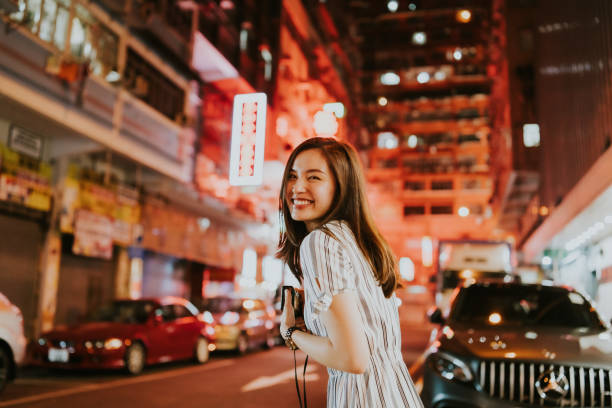 young cheerful asian woman looking back in front of the neon lights of hong kong  on the streets at night while holding her camera - hong kong china city night imagens e fotografias de stock