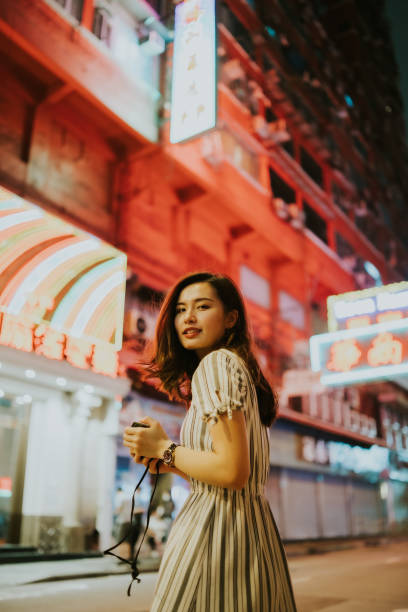 istock Young Asian woman smiling and looking back while holding her camera and standing in front of the street with neon light shop signs and building 1425538603