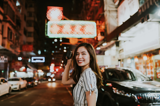 Young Asian woman holding her camera towards the street with neon lights and traditional buildings while looking back at camera, in the city of Hong Kong. An Asian girl tourist using her camera to photograph around the busy streets with traditional neon signs in Hong Kong at night. A portrait of a happy female traveler standing in front of a busy street with shops and neon boards, smiling towards camera and discovering the urban cityscape whiile taking photos in the evening.