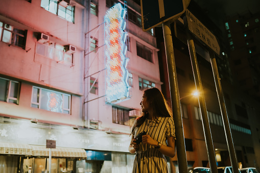 Young smiling Asian woman holding her camera in hand while she looks at the view at night with a background of neon lights,cars and old buildings in the city of Hong Kong. A cheerful Asian girl enjoying the view around the city while taking pictures using the camera, during night-time while she stands in front of a street pole/road sign. A portrait of a happy female traveler relaxing and discovering the streets of a buzzing city in the evening while taking pictures.