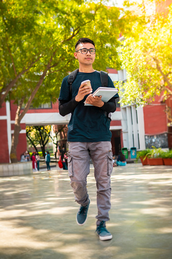 Low angle image of happy east Asian teenager boy student walking at the university campus by holding books and coffee mug in hand.