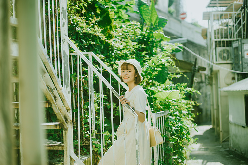Young smiling Asian Woman looking at the camera for a photo under the warm sunlight while walking the staircase in an old fishing village as she explores the life in Hong Kong Island in summer holiday. 
Cheerful Girl holding the handrail as she walks up the stairs while enjoying the nature view with trees and old houses in a narrow street, to relax in a sunny day inside a rural village. 
A portrait of a happy female traveler standing on the staircase near the quiet non-urban area of a city during her vacation tour.