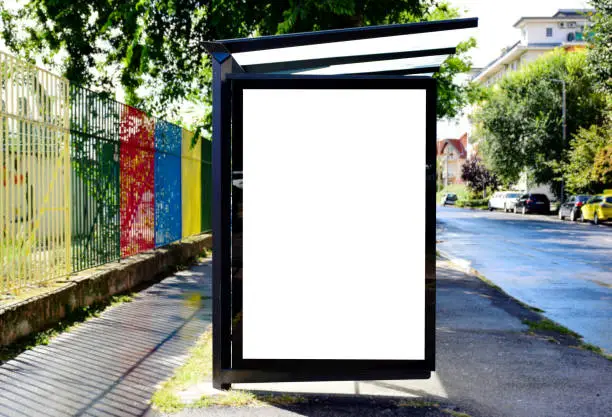 empty milky white poster ad and advertising display glass and light box. image composite of bus shelter at a bus stop. glass and aluminum frame structure. street perspective with colorful metal fence and blue bus. green background.