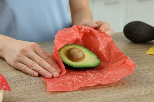Woman packing half of avocado into beeswax food wrap at wooden table, closeup
