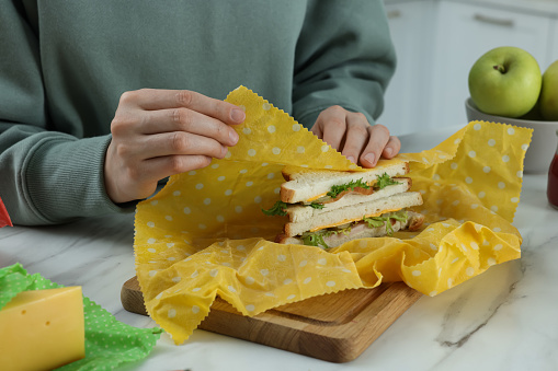 Woman packing tasty sandwich into beeswax food wrap at white marble table indoors, closeup